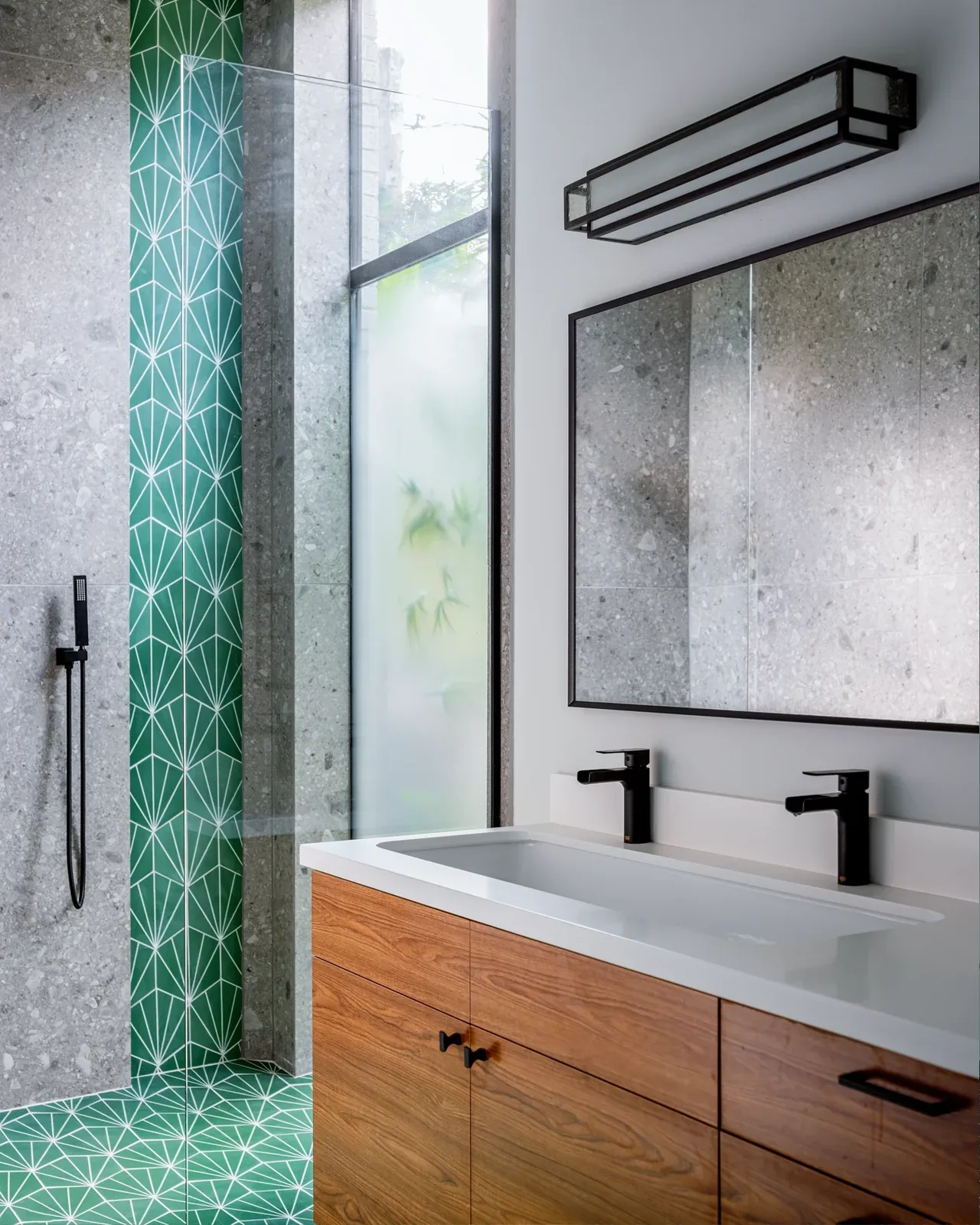 Bathroom featuring a double vanity with black faucets, green geometric tile, and a frosted window next to a shower area.