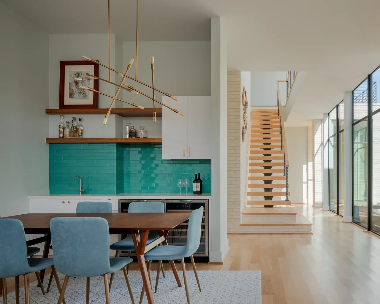 Open dining space with modern kitchen featuring teal backsplash, wooden table, and a staircase leading to upper floor in Windemere Residence.