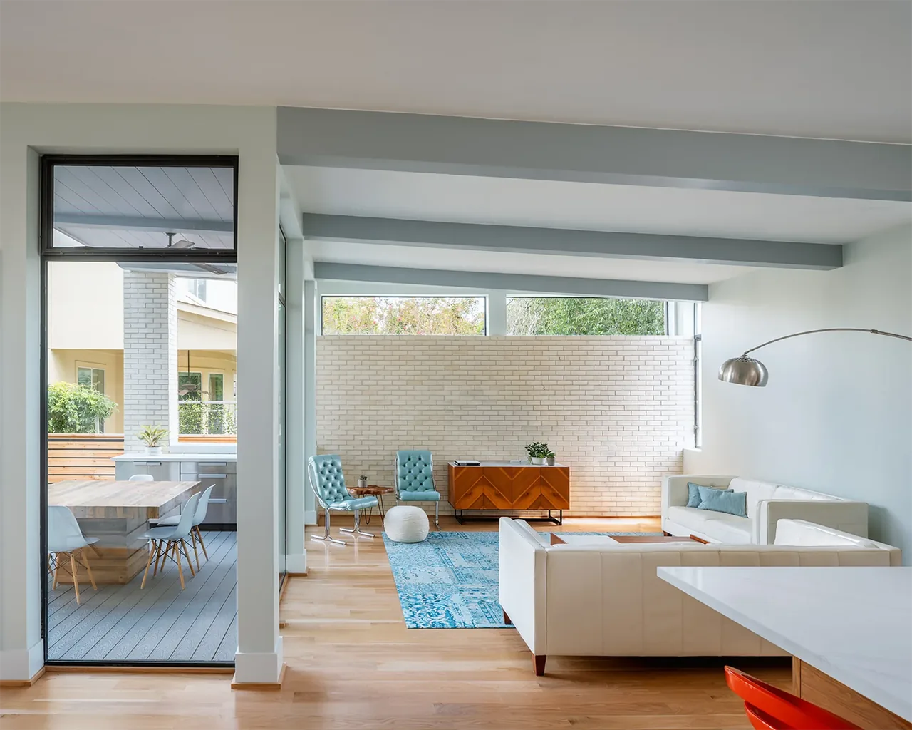 The Windemere Residence living area features a white brick wall, wood floor, blue accent furniture, and a view to the outdoor dining space.
