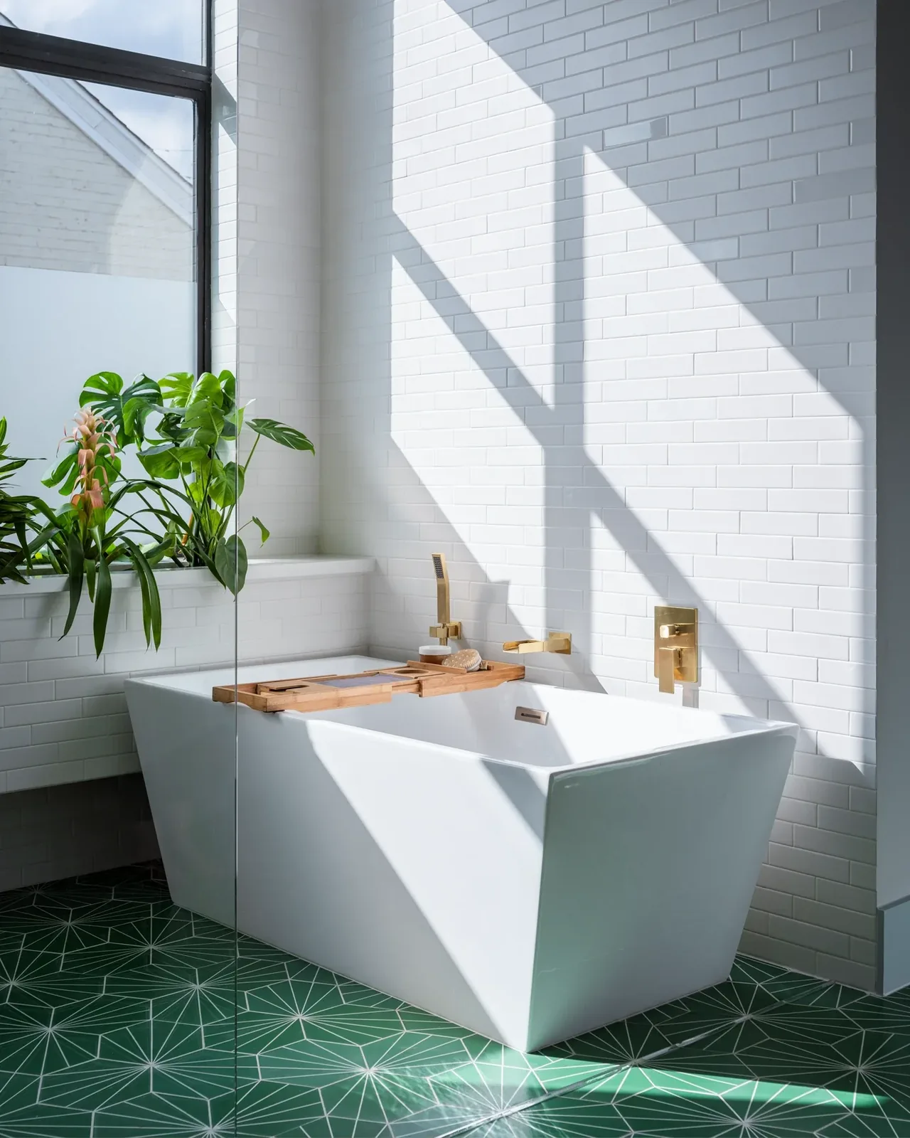 Bright bathroom corner with a white freestanding tub, brass faucet and shower handle, green geometric tile floor, and a large window sill planter.