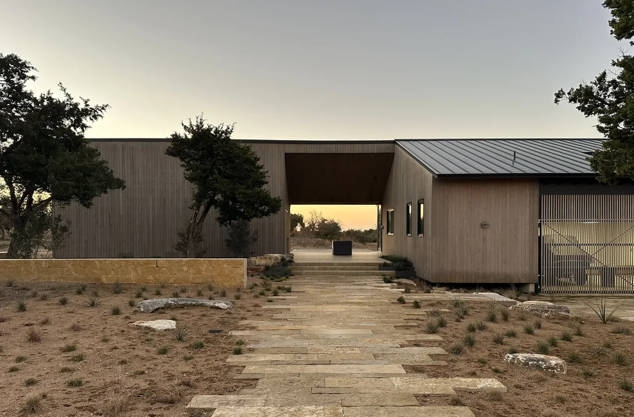 Wimberley Residence features vertical wood siding and a covered passage beneath a flat roof, accented by large rectangular windows.