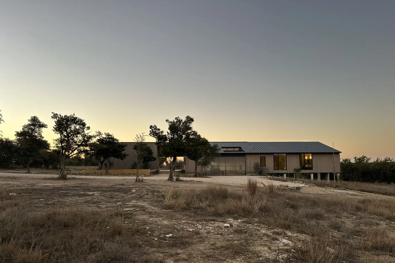 The Wimberley Residence features an elevated structure with vertical wood siding and large windows set in a dry grass landscape at dusk.