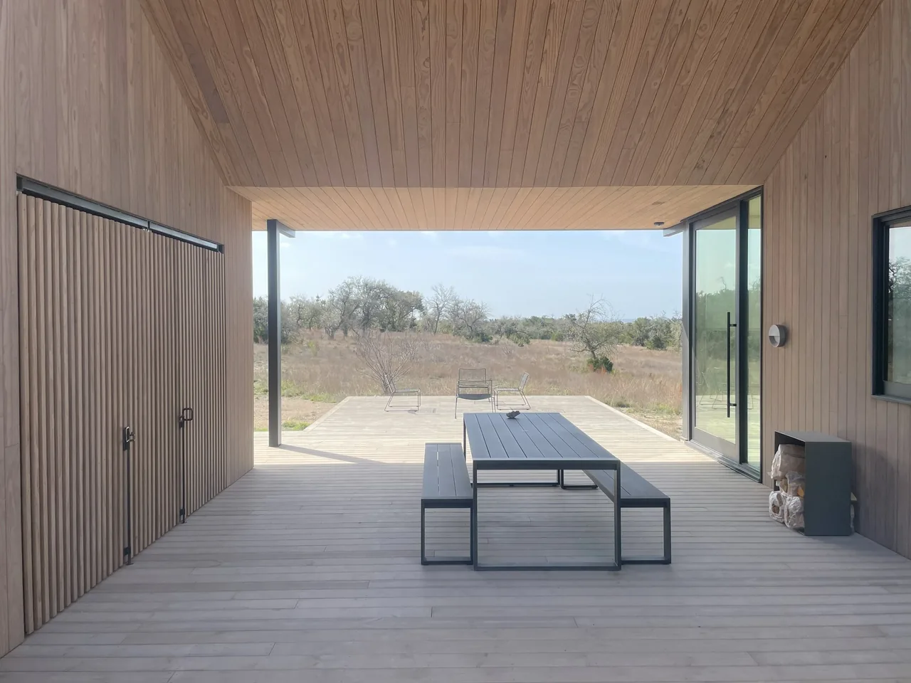 Covered patio with vertical wood siding and a wood plank ceiling extends to an open deck overlooking a dry grass landscape.