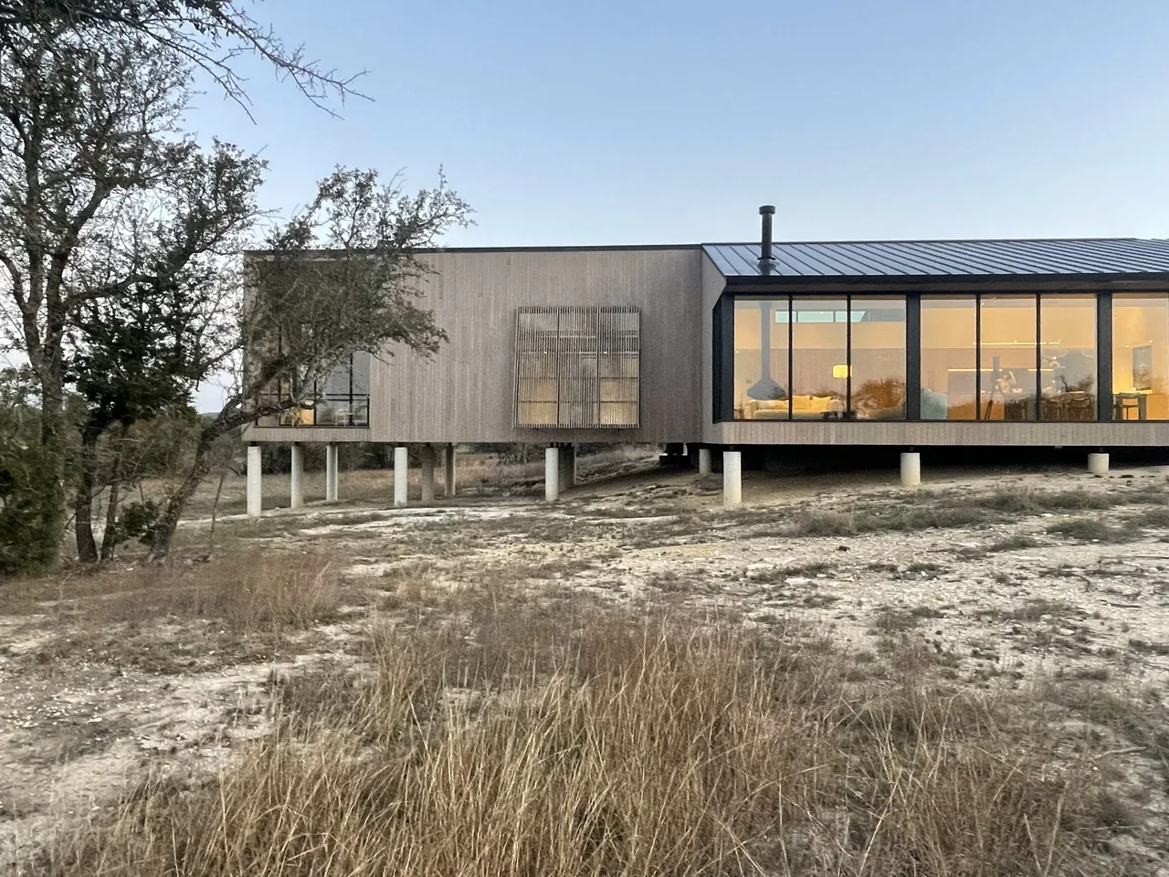 Wimberley Residence features an elevated structure with vertical siding and large rectangular windows in a dry grass landscape.