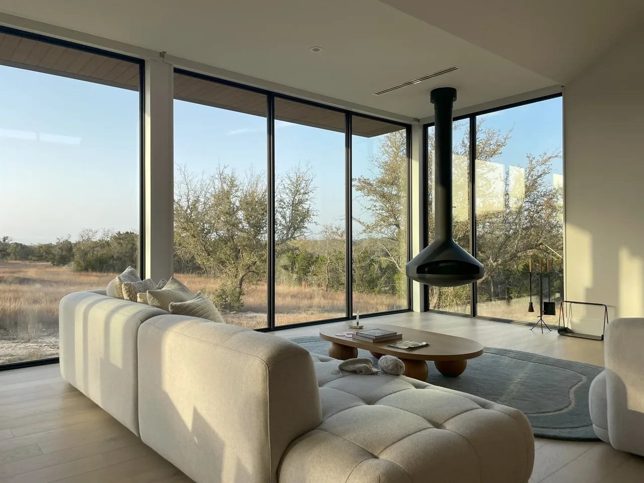 Living room with floor-to-ceiling corner windows framing a dry grass landscape, a suspended black fireplace, and modern cream upholstery seating.