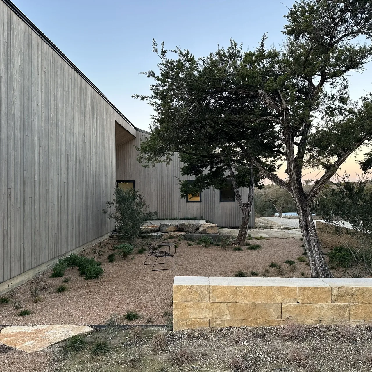 Wimberley Residence features vertical wood siding and natural stone steps in a dry grass landscape with shrubs.