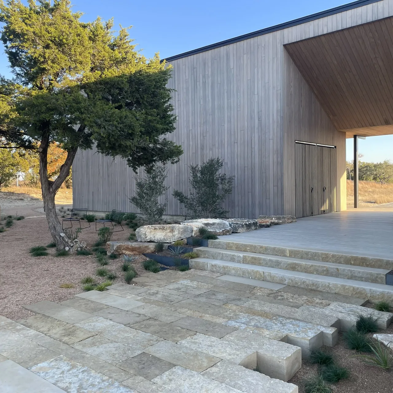 Wimberley Residence features vertical wood siding, stone steps, and a covered patio area in a dry grass landscape.