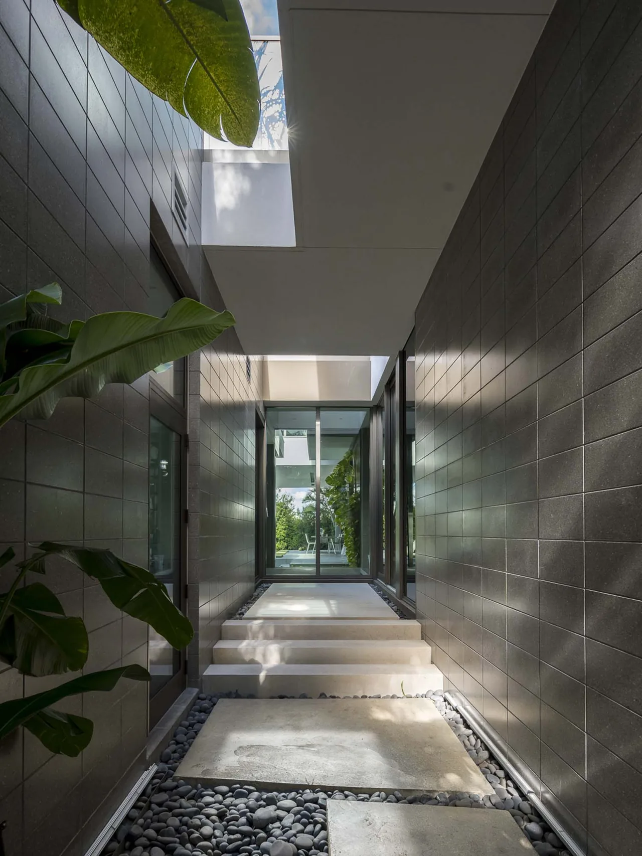 Narrow corridor in Intracoastal Residence features dark tile walls, concrete stepping stones over pebbles, and overhead sky openings.