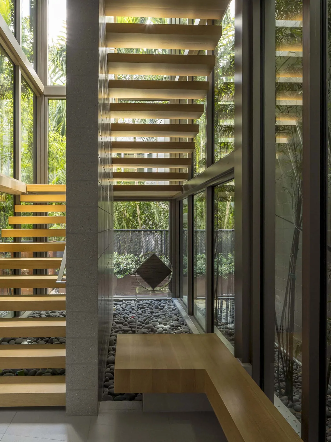A modern wooden floating staircase next to floor-to-ceiling glass windows with a view of a pebble garden and greenery.