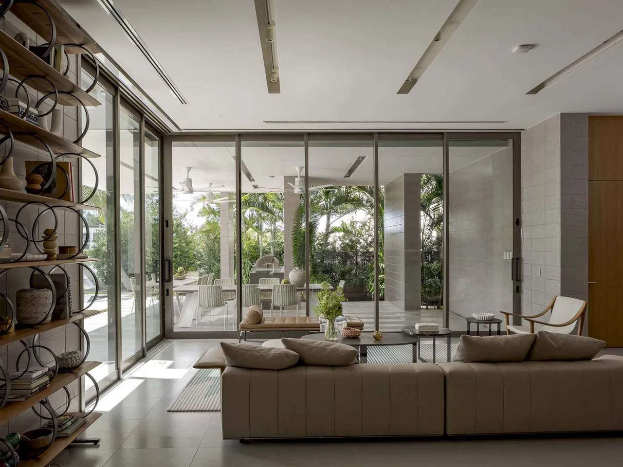 The living room features floor-to-ceiling sliding glass doors that lead to a covered outdoor patio with lush tropical landscaping.