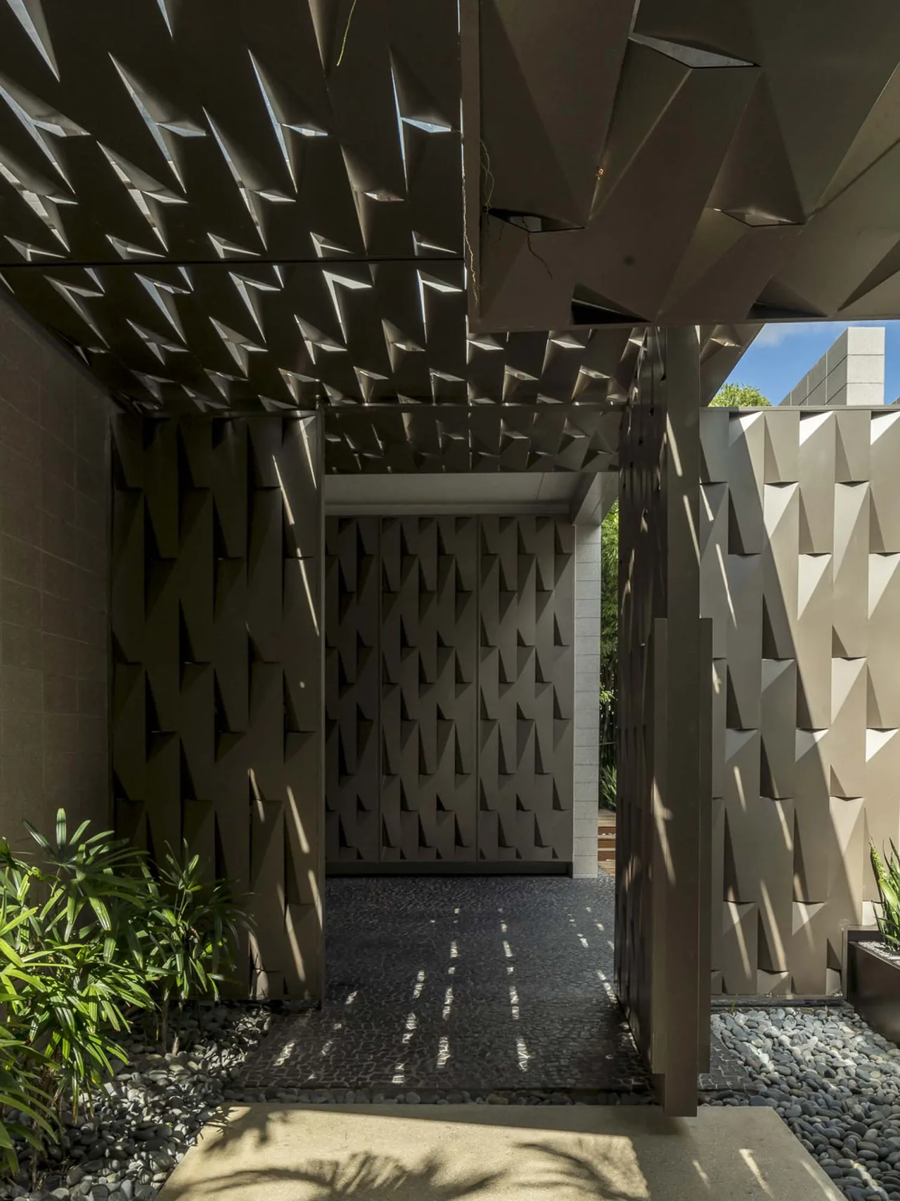Geometric metal canopy creates patterned shadows over a corridor with textured walls and stone pebble flooring at Intracoastal Residence.