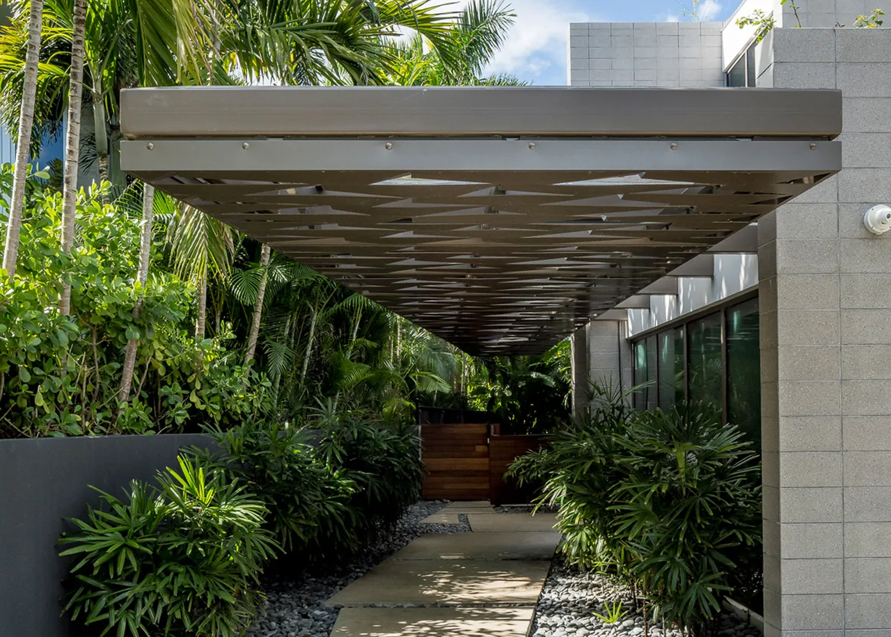 View of a shaded outdoor walkway with a decorative metal canopy and lush green plants on both sides.