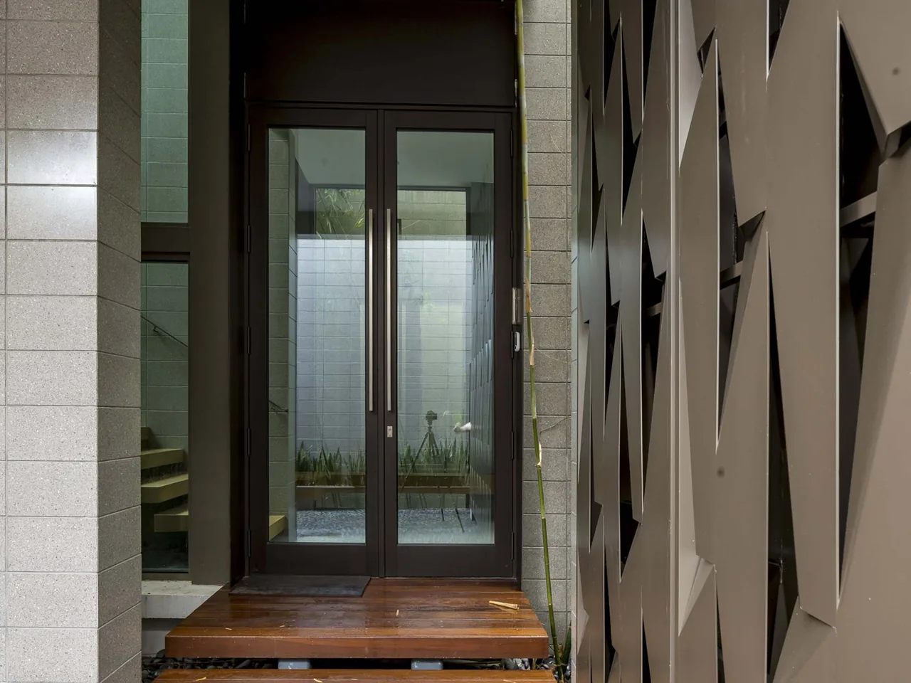 Entrance corridor features textured wall panels, natural stone pebbles on the ground, and a geometric metal canopy casting angular shadows.