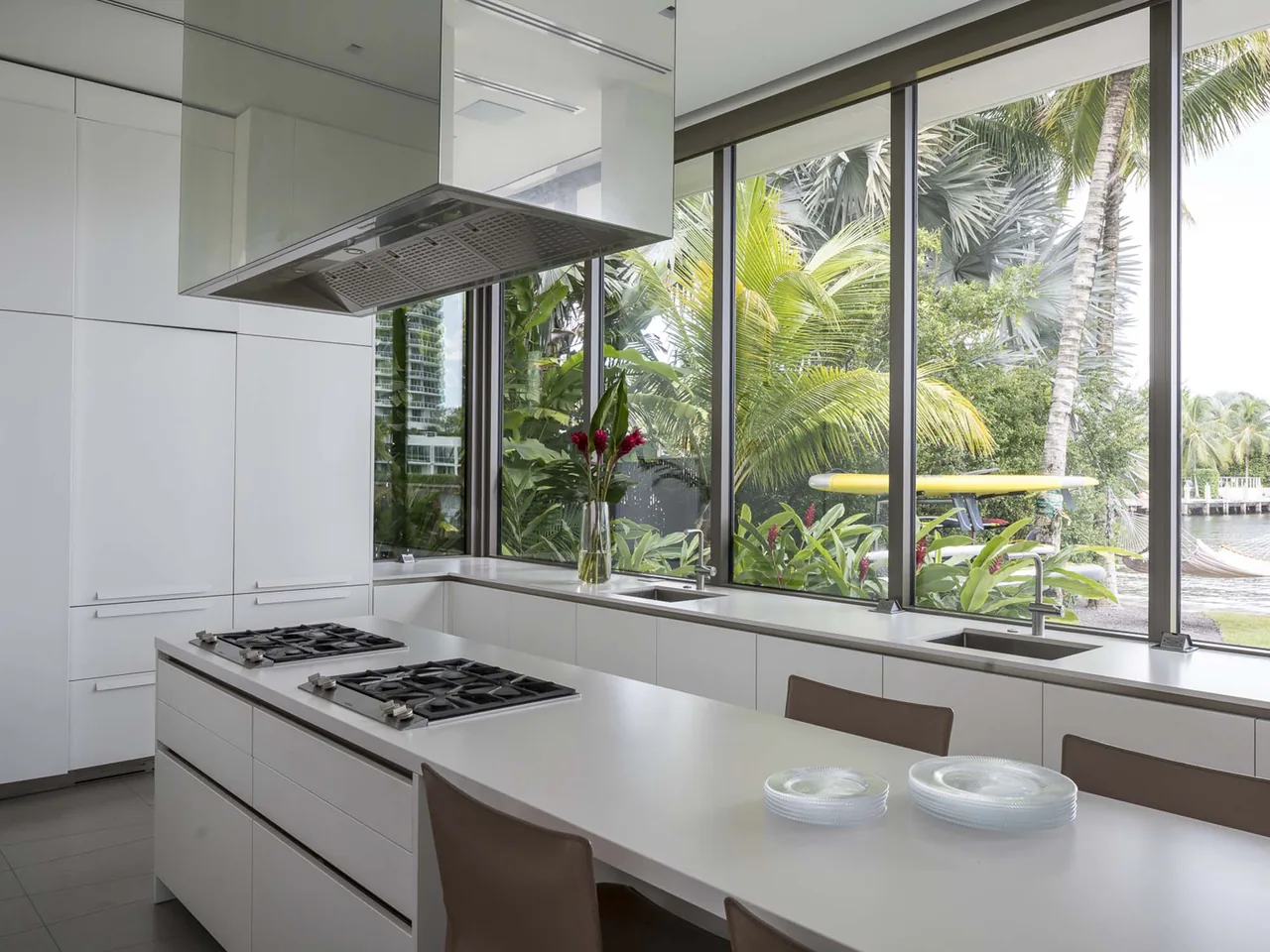 The Intracoastal Residence kitchen features a white island with gas cooktops and large windows overlooking lush tropical plants.