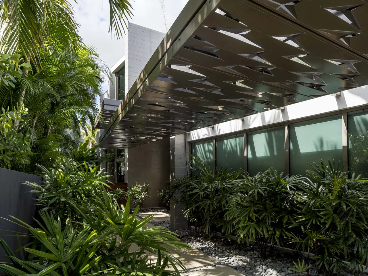 The Intracoastal Residence features a metal canopy corridor flanked by tropical plants and natural stone pebbles along the pathway.