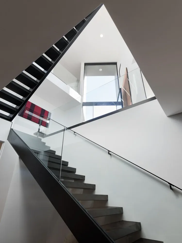 Modern staircase with black treads and glass railing illuminated by natural light from a large upper window.
