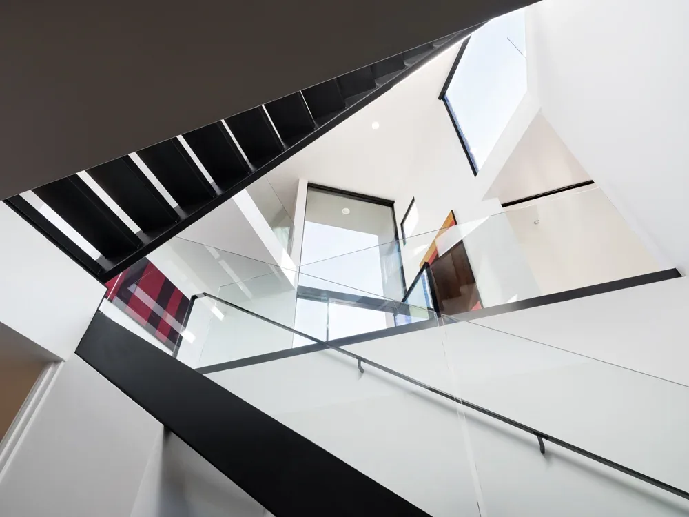 Floating black stairs with glass railings ascend beside large windows in the bright Neptune House interior.