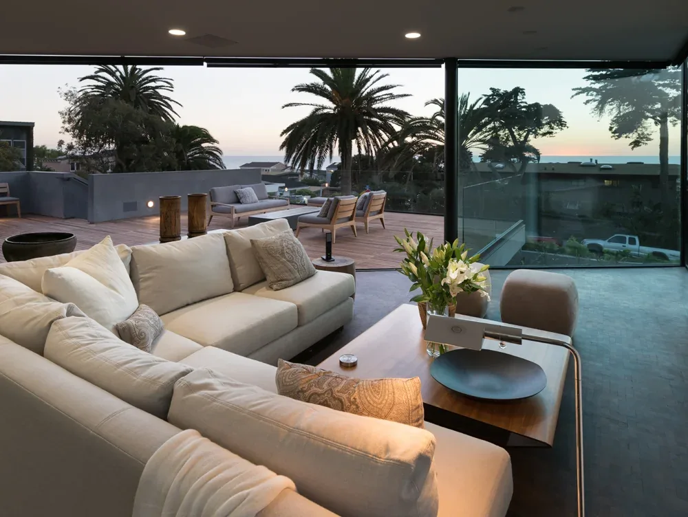 Living room with a beige sectional sofa and coffee table, seamlessly connecting to an outdoor deck with seating and ocean views.