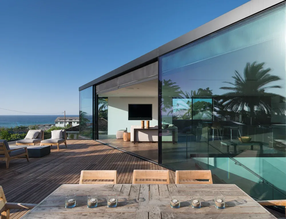 Neptune House features a wooden outdoor deck with lounge chairs and large glass walls connecting indoor and outdoor spaces.