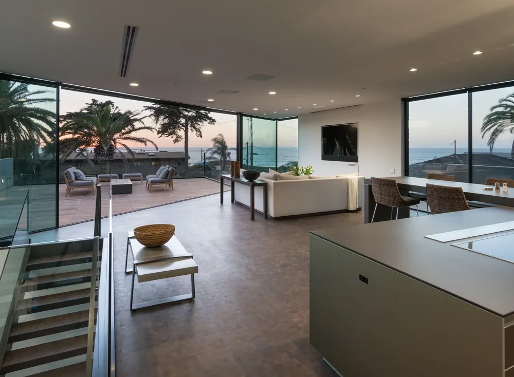 Living room and kitchen area feature floor-to-ceiling glass walls with ocean views and access to an outdoor deck with lounge seating.