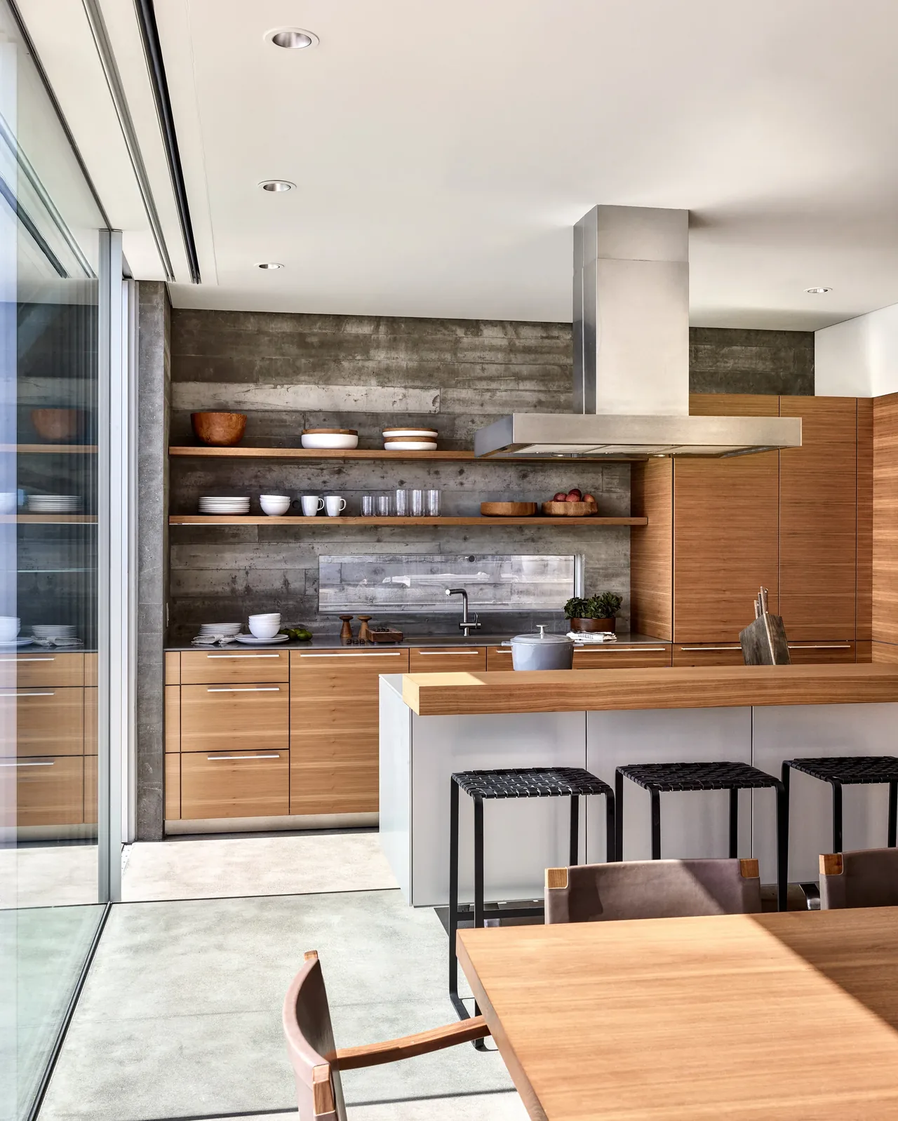 The Levine House kitchen features horizontal wood cabinetry and a stainless steel range hood against a textured concrete wall.