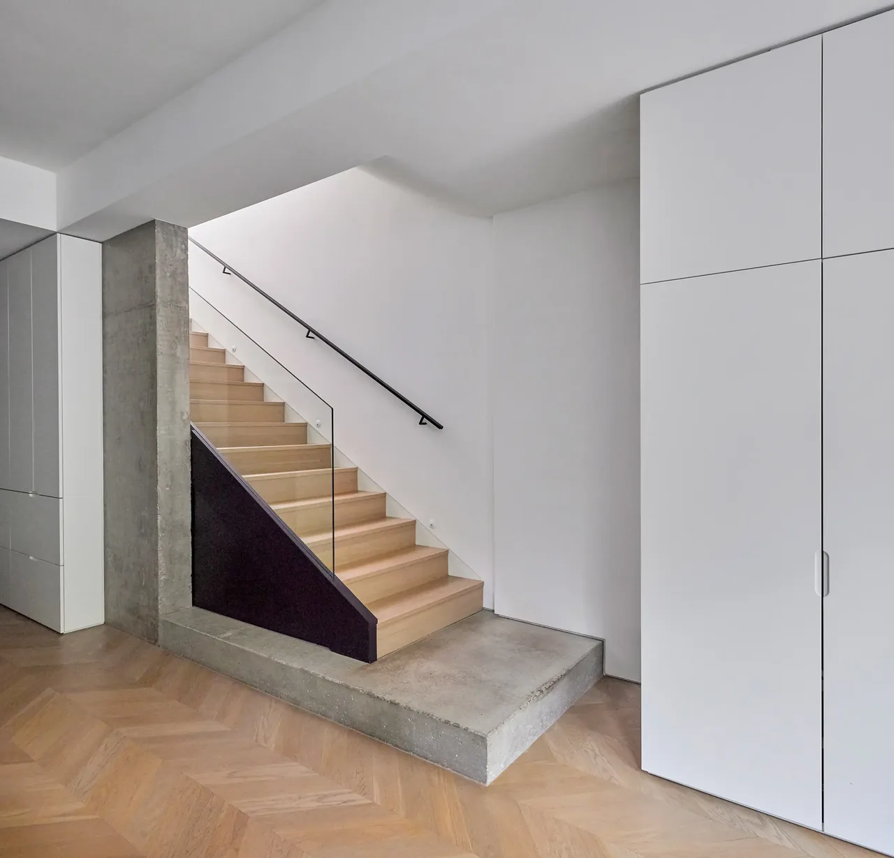 Staircase features light wood steps, glass railing, black handrail, and a concrete platform base.