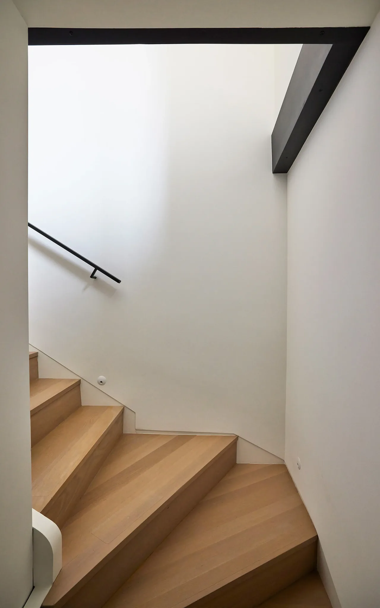 A modern wooden staircase with white walls and a black handrail in a minimalist setting.