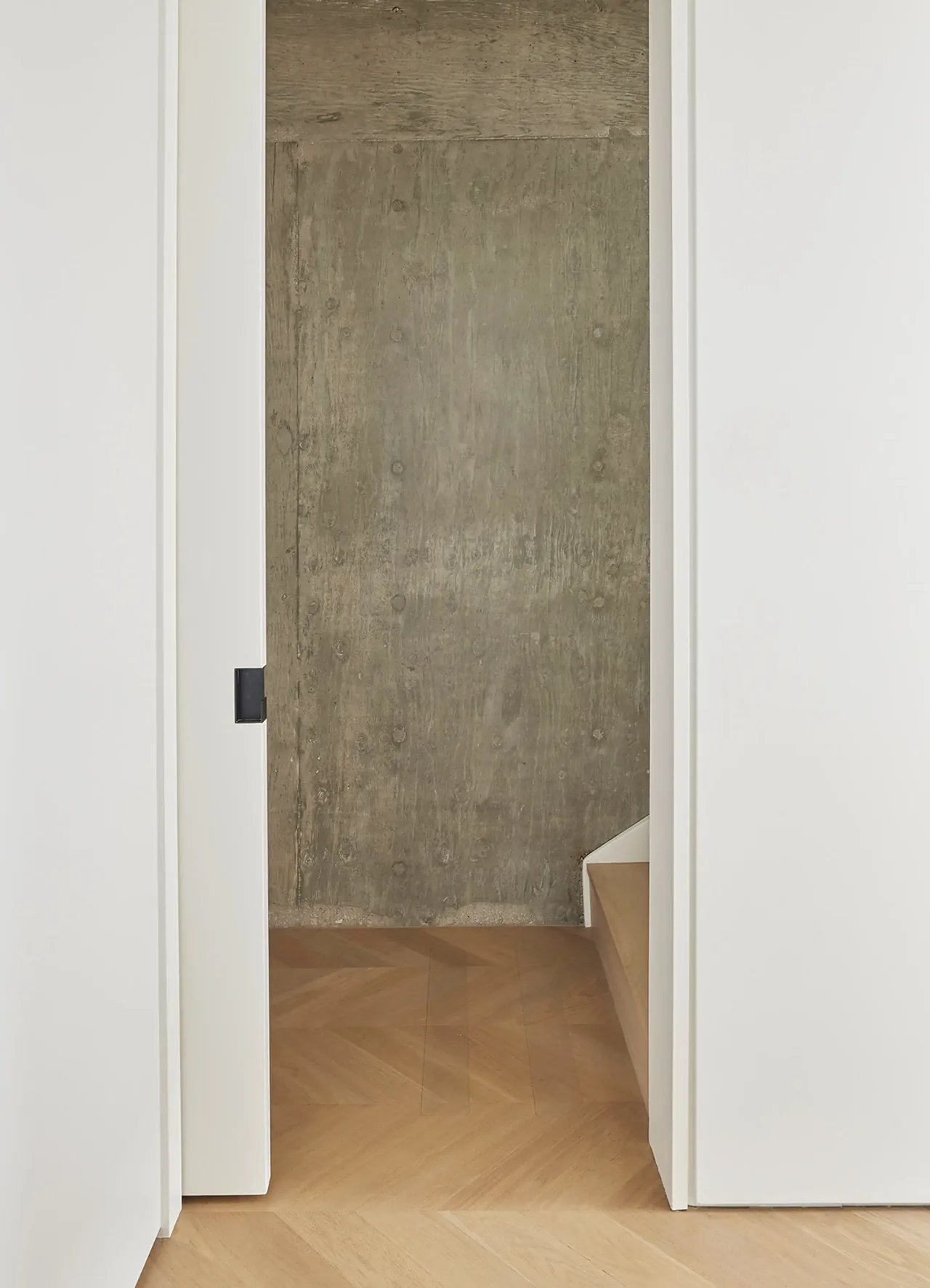 Narrow hallway leading to stairs with herringbone wood floor and a concrete feature wall.
