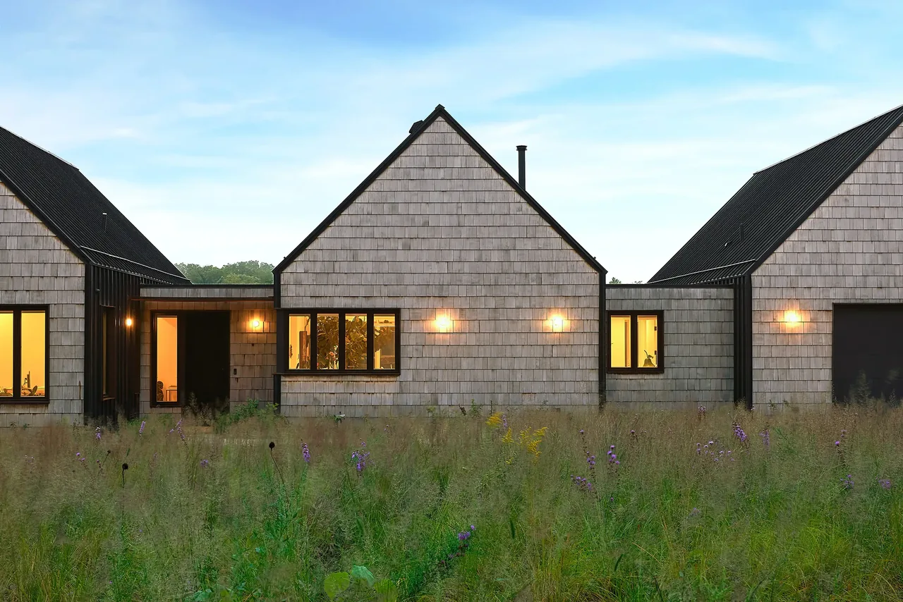 Lake Geneva House features connected pavilions with beige shingle siding and contrasting black metal roofs, illuminated in evening light.