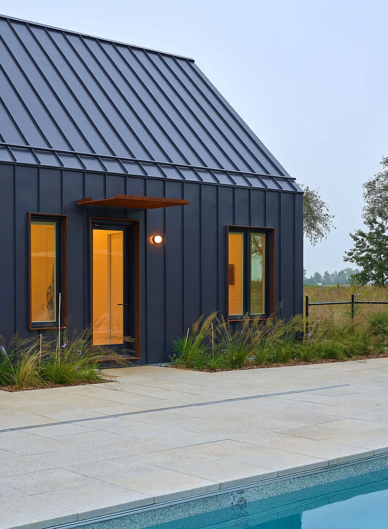 Side view of Lake Geneva House featuring black metal siding, tall rectangular windows, and poolside paving.