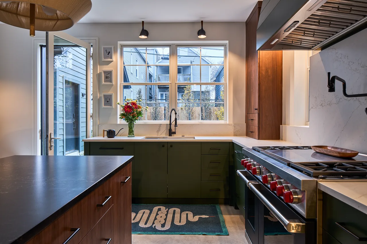 The Mozart House kitchen features green cabinetry, a black countertop island, stainless steel range with red knobs, and a view through the window.