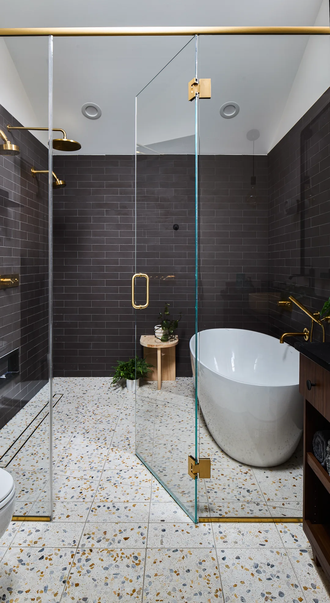 This bathroom features terrazzo tile flooring, a freestanding tub, and dark tile walls with brass shower fixtures.
