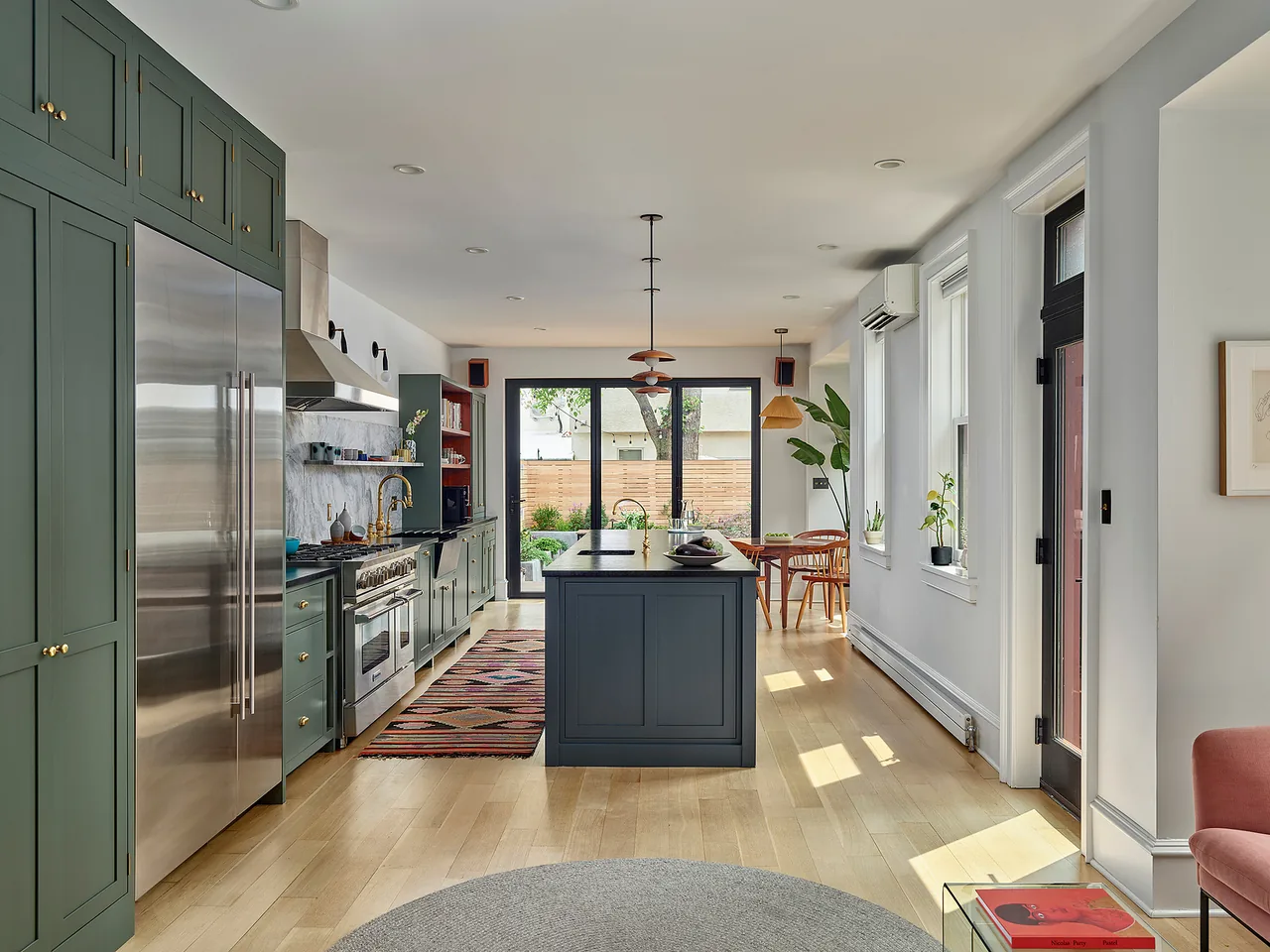 Kitchen with green cabinets, large island, wood dining chairs, and folding glass door to garden patio in the Fishtown Rowhouse Addition.