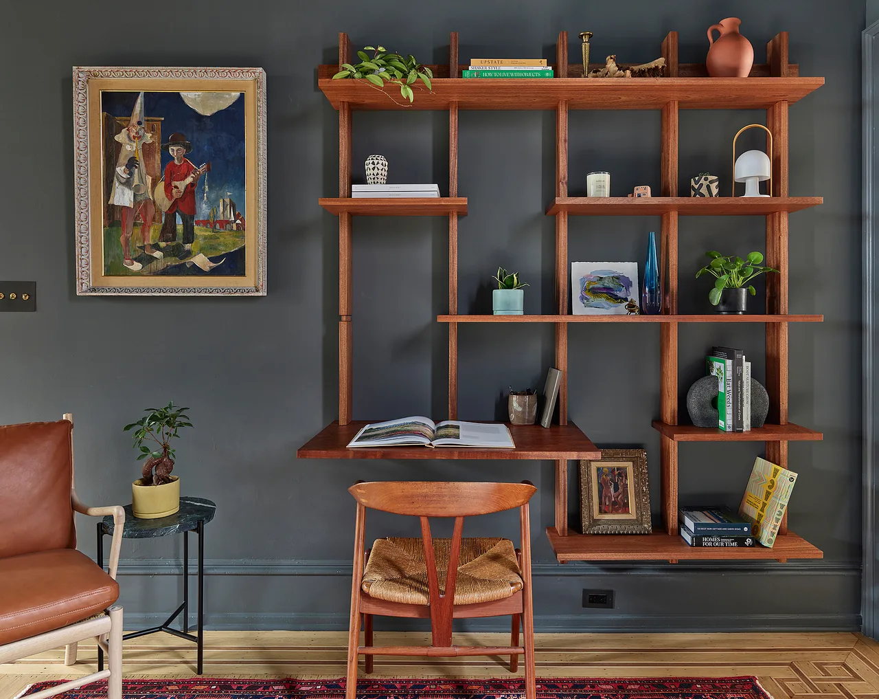 Wall-mounted wooden shelving with an integrated desk and a leather chair in a gray-painted room with artwork.