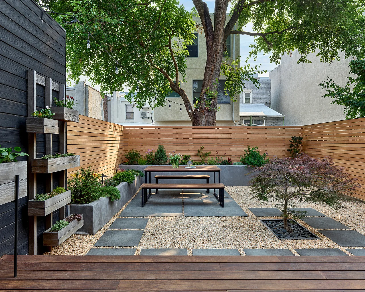 Backyard patio with wood fencing, concrete planters, and a dining set beneath string lights and a large tree canopy.