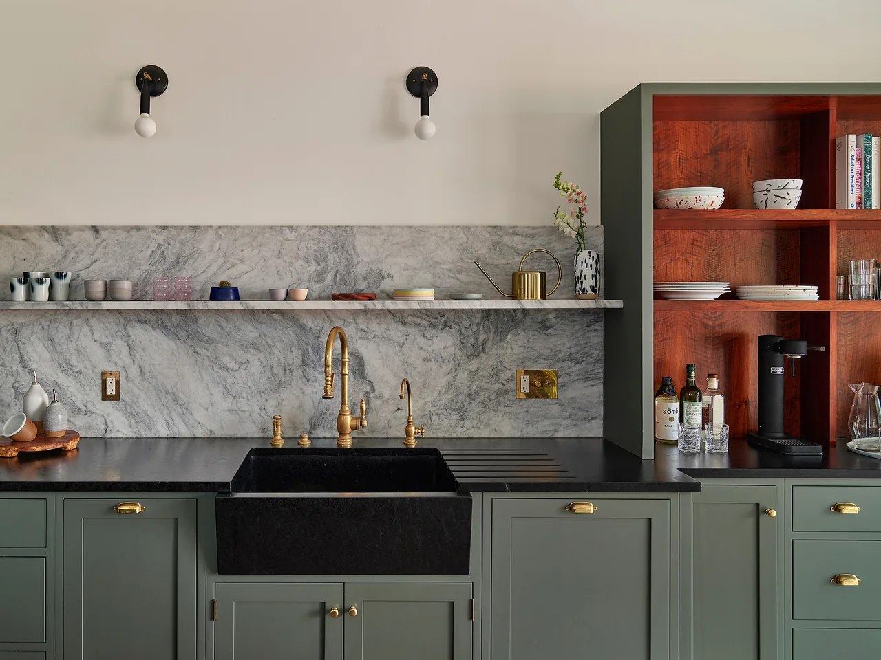 Kitchen interior featuring green cabinetry, a black farmhouse sink, brass fixtures, and marble backsplash with open wooden shelving.