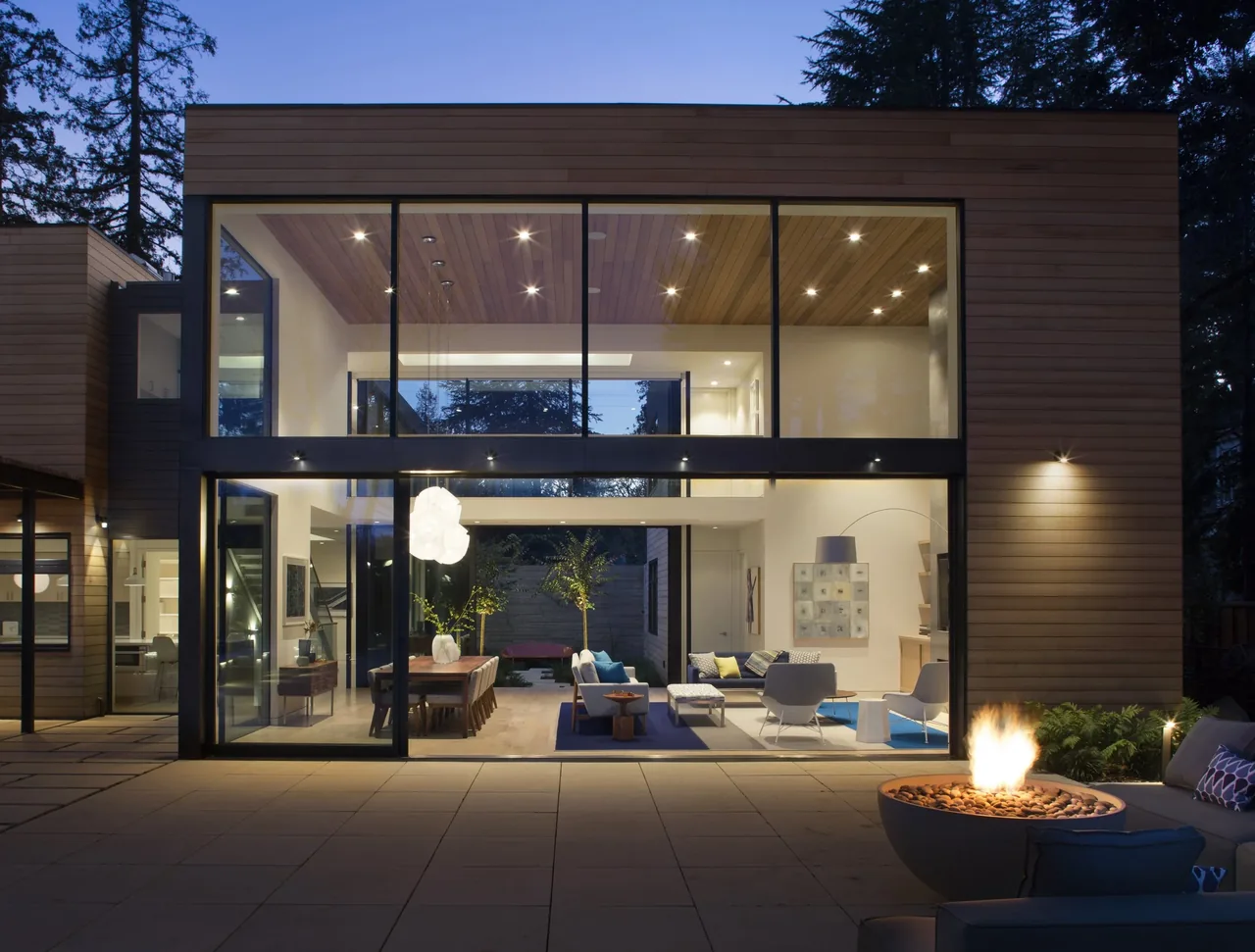 Hermosa Residence features large floor-to-ceiling glazing and horizontal wood siding at night with an outdoor fire pit.