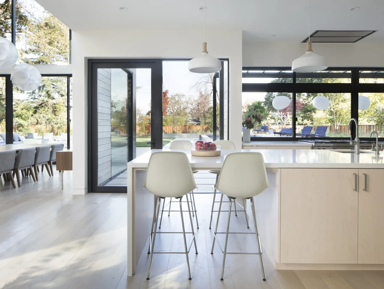 Kitchen area with light wood cabinetry, white pendant lights, and large black-framed windows providing views to the outdoor patio and garden.