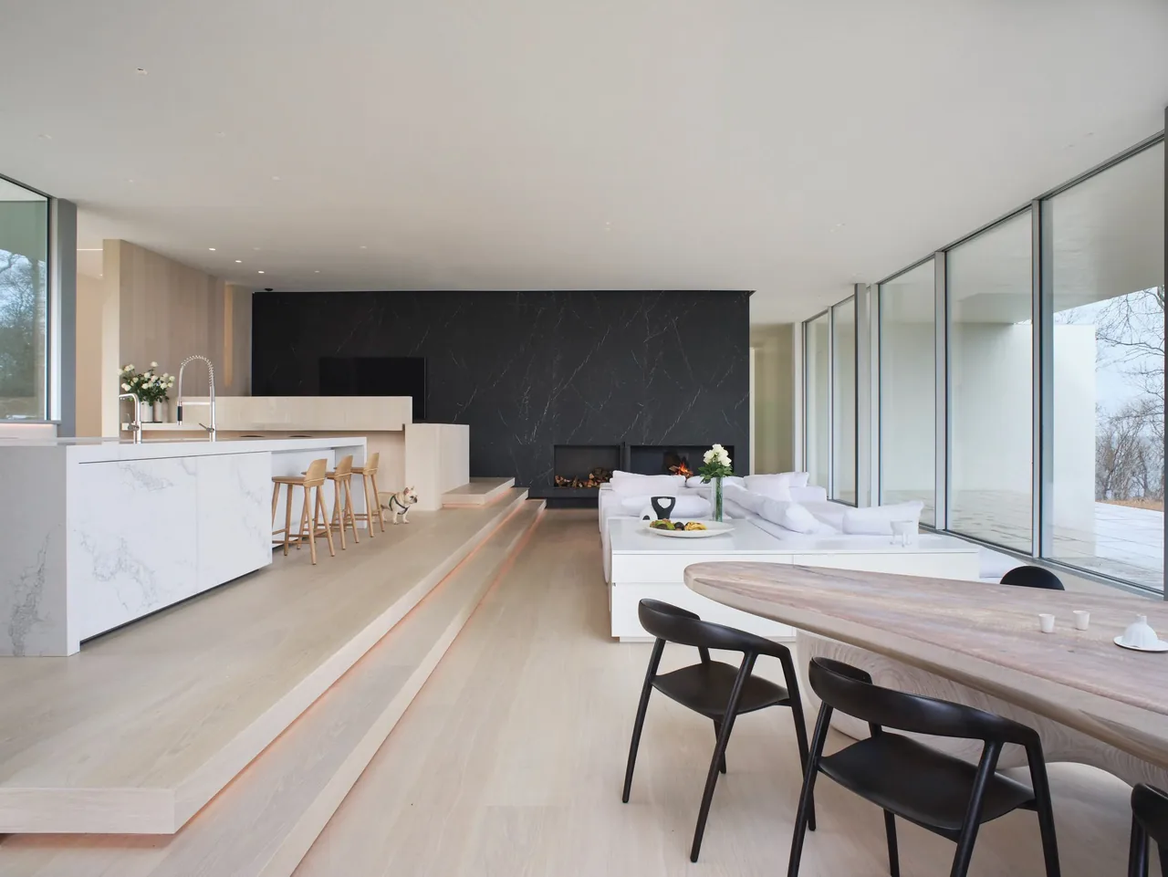 Open living and kitchen space with a black marble fireplace wall and large floor-to-ceiling glass windows overlooking a snowy landscape.