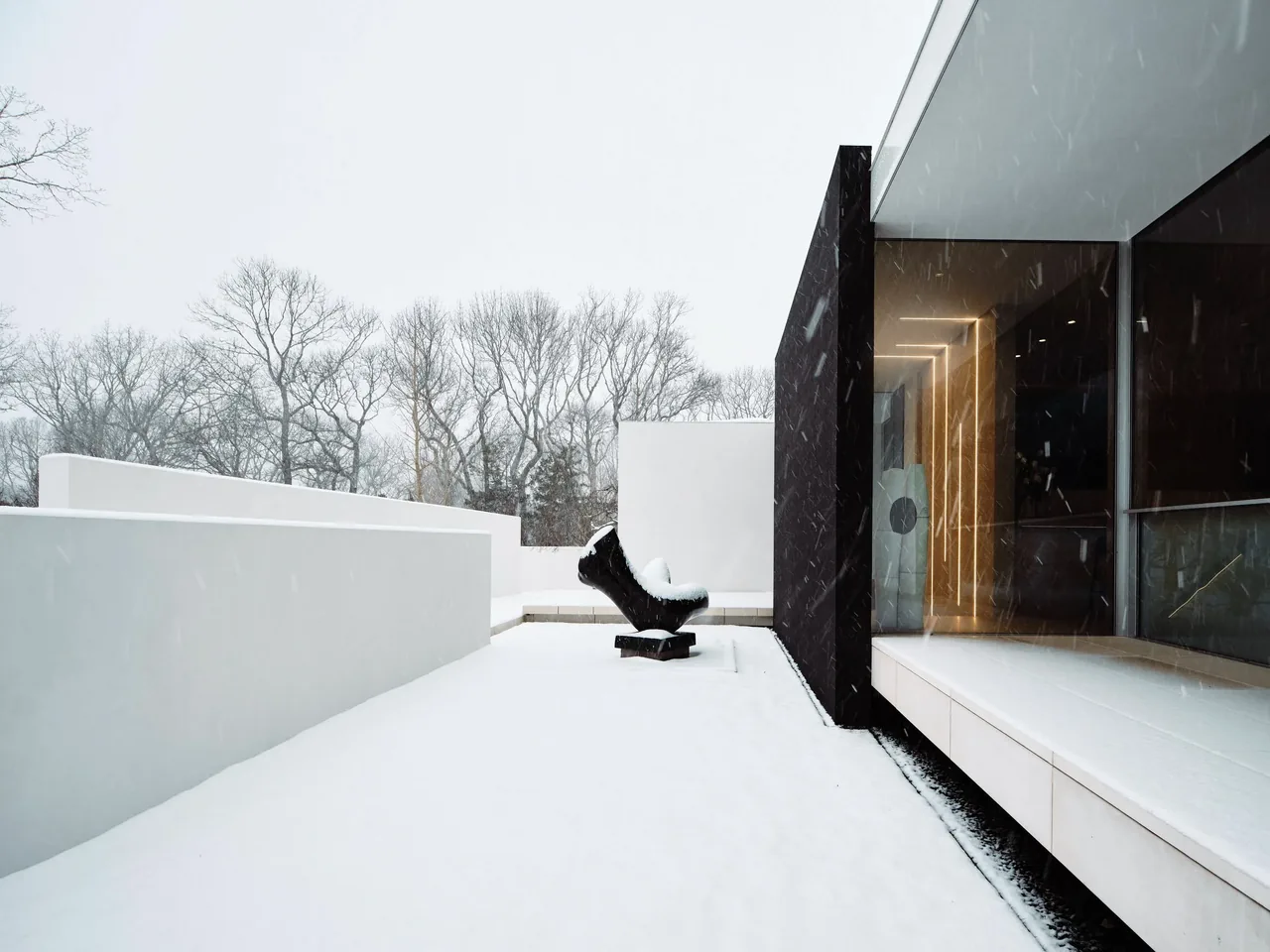 Modern minimalist exterior patio covered in snow with a dark abstract sculpture amid leafless trees.