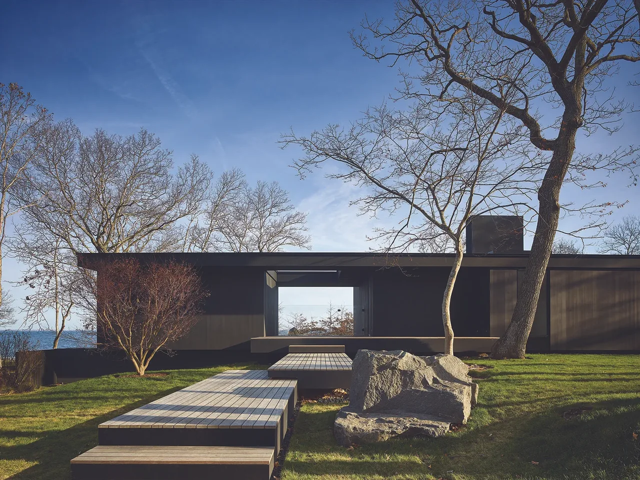 Modern black exterior house with a flat roof and a walkway leading through a lawn with trees and large rocks.