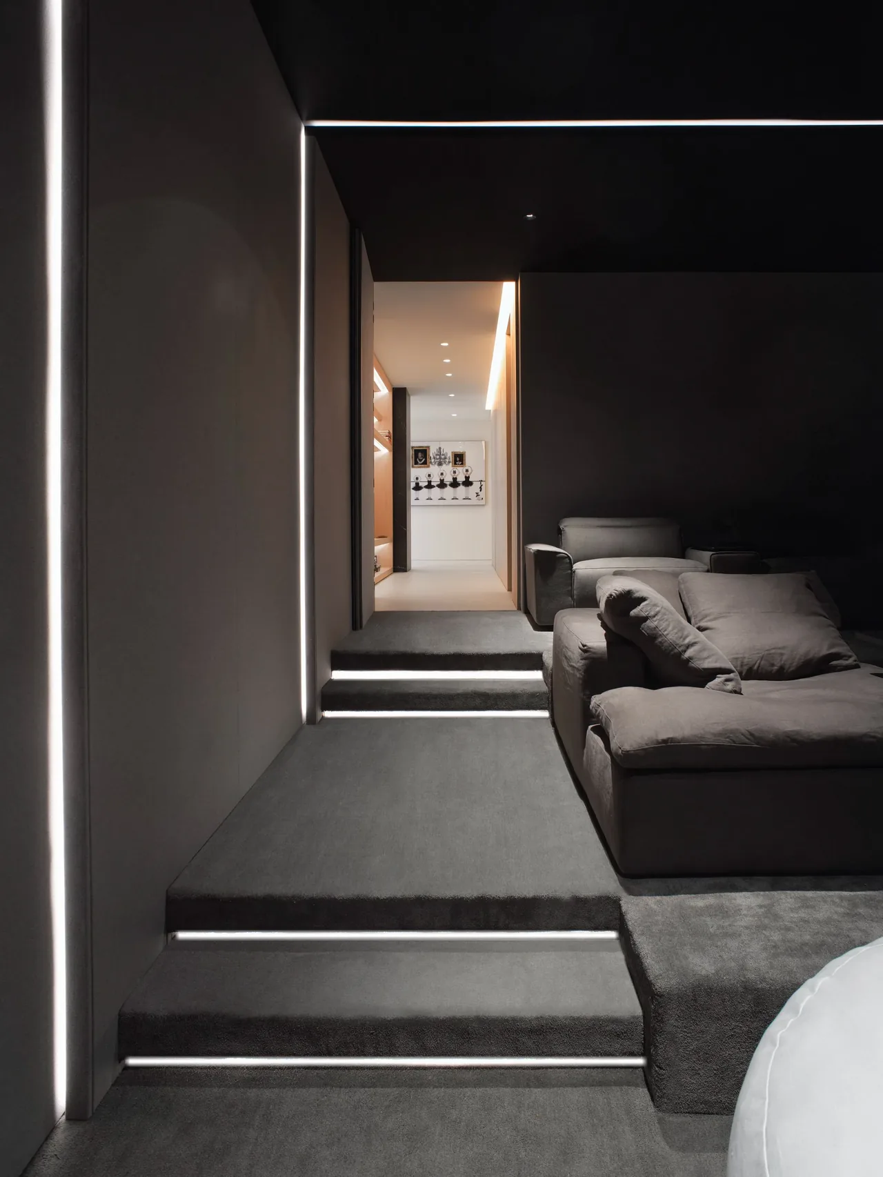 Living area with gray cushioned furniture, carpeted stairs with linear lights, and a lighted hallway leading to built-in shelving.