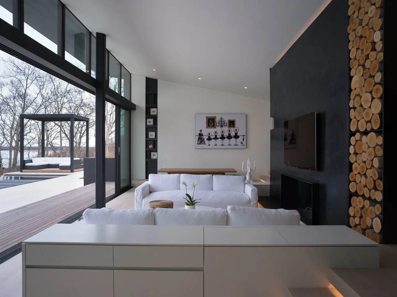Living room with floor-to-ceiling glazing, black fireplace, and a wall stacked with round wood logs next to the TV.