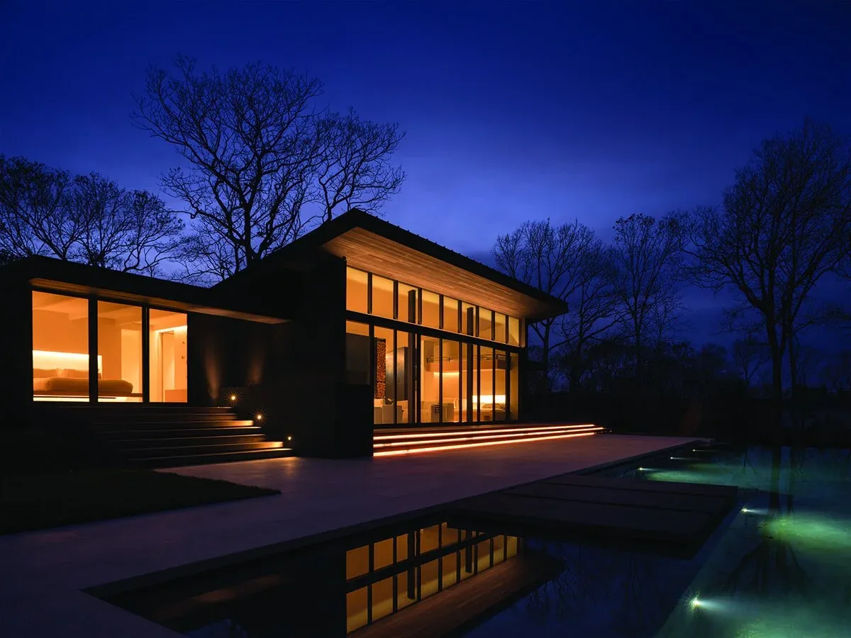 The Lighthouse & Treehouse project features a modern home with floor-to-ceiling glazing and illuminated exterior stairs by the pool at night.