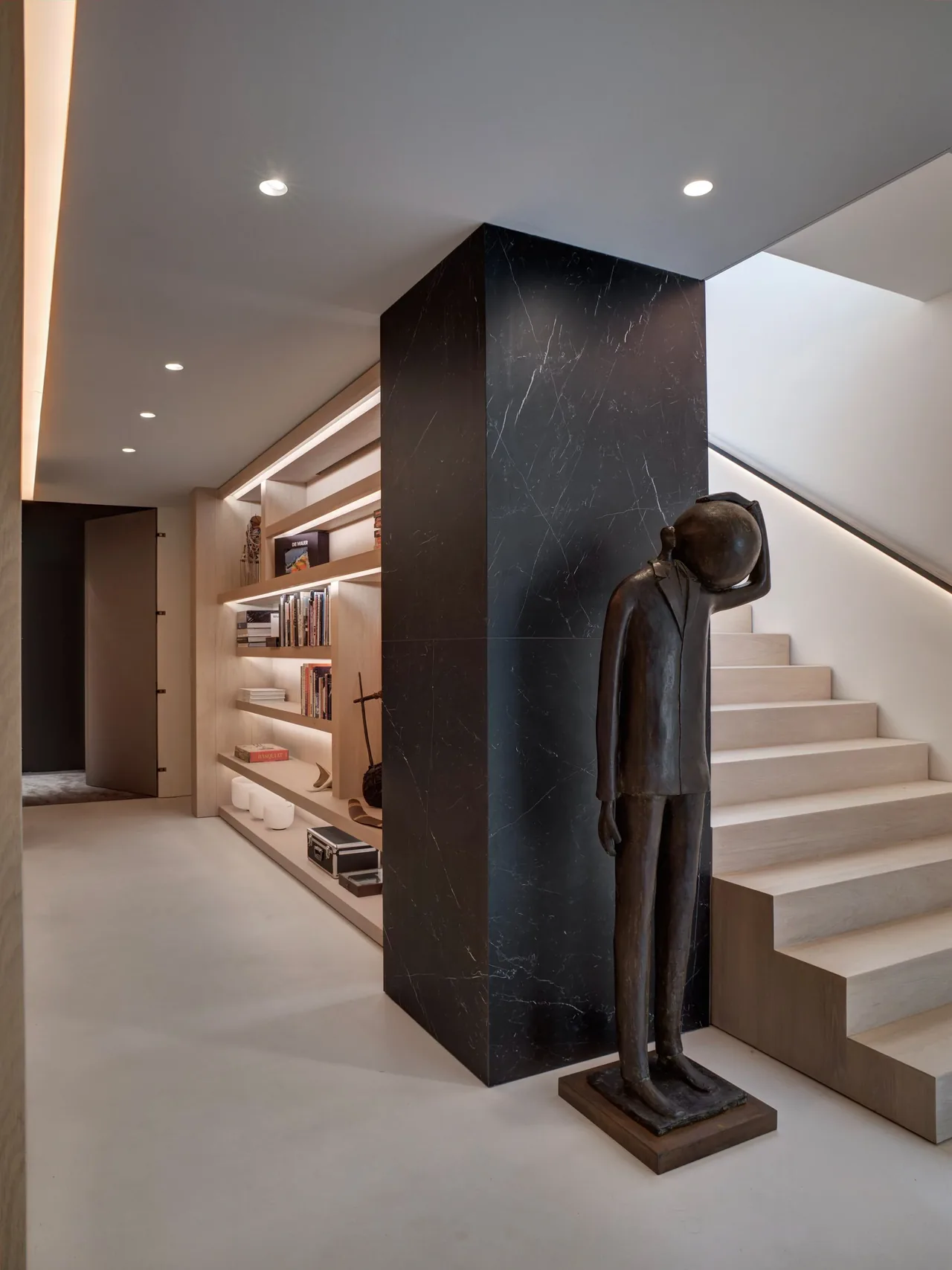 Hallway featuring a black marble column, light wood built-in shelves, and a bronze statue near a light wood staircase.
