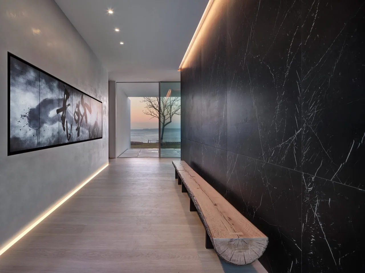 Entry hallway features a long, raw wooden bench against veined black marble and abstract art, leading to a glass door overlooking a leafless oak and waterfront at sunset.