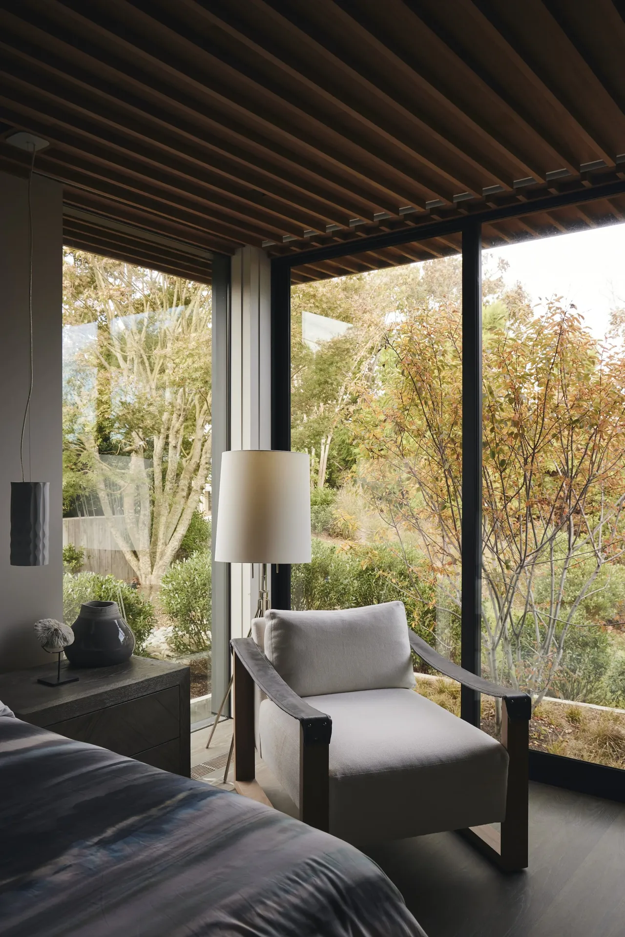 Bedroom corner with a wooden slat ceiling, floor-to-ceiling glazing, and a view of garden plants.