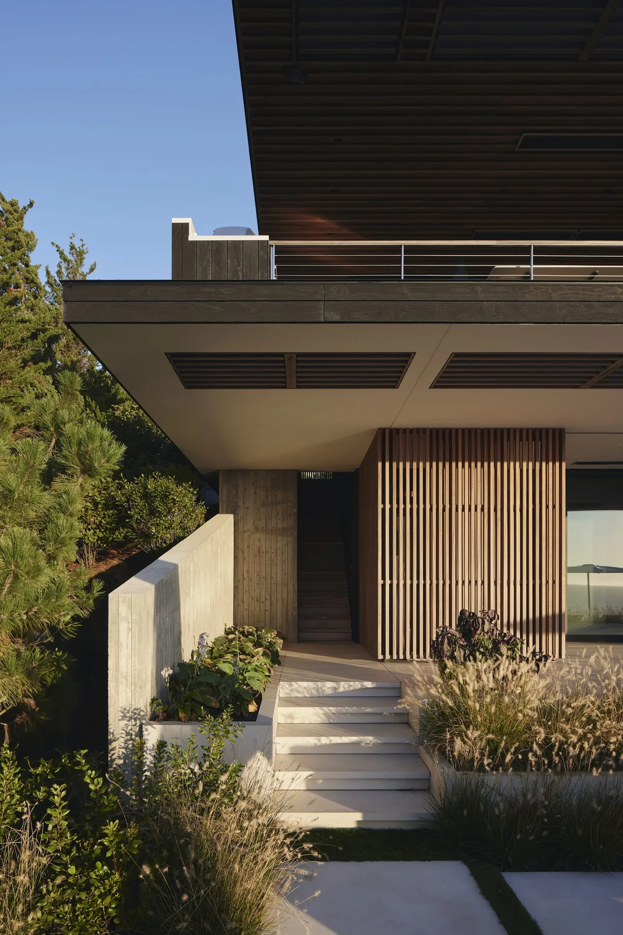 Modern beachfront home entrance with vertical wood slat screen and concrete stairs framed by plants.