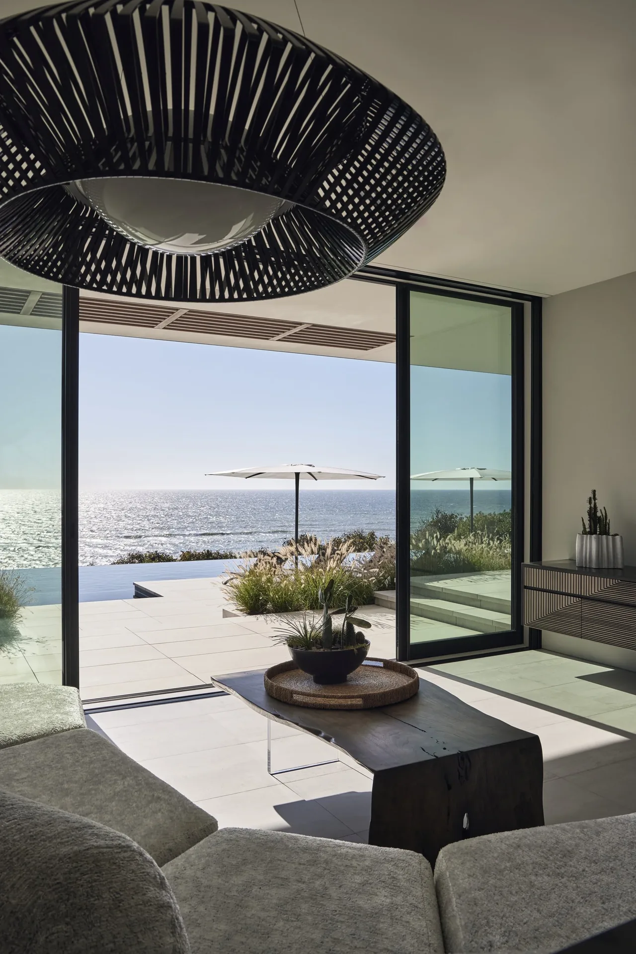 Living room with large floor-to-ceiling sliding glass doors overlooking an ocean and patio area with plants and an umbrella.