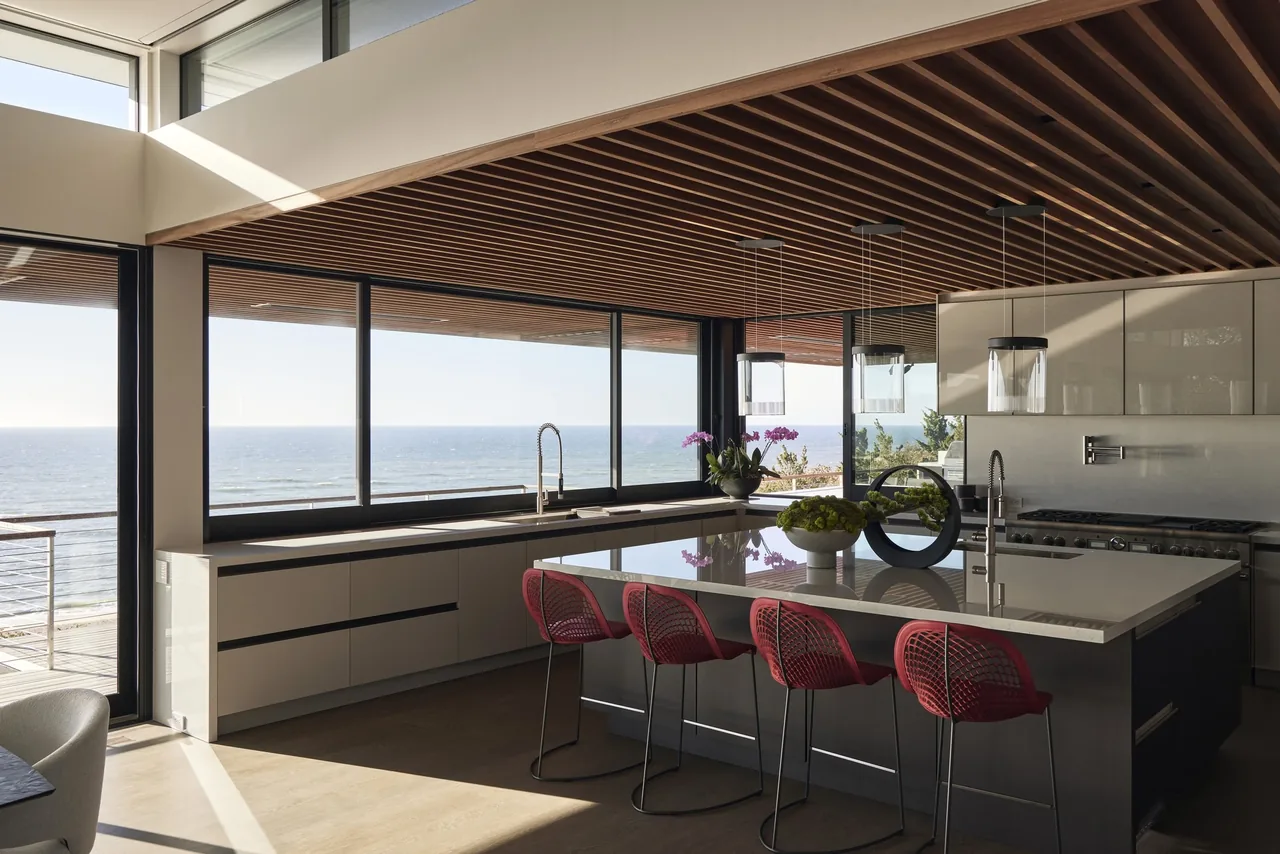 Ocean Bluff kitchen features a large island and expansive windows overlooking the ocean, with a wood slat ceiling and pendant lighting.
